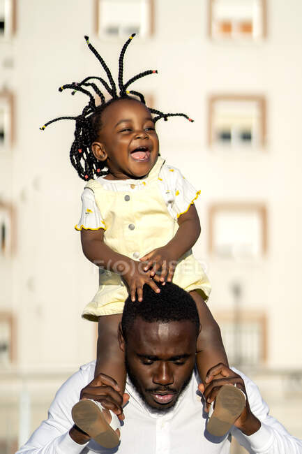 Веселий афроамериканець у сорочці, який носить маленьку дочку за плечима і стрибає, проводячи разом час на вулиці під сонячним світлом. — стокове фото
