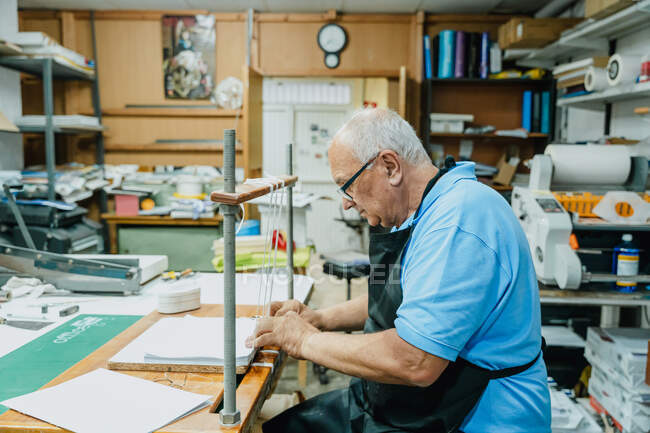 Vista lateral do artesão masculino sênior atencioso em avental e óculos amarrando fitas em placa de madeira antes de trabalhar na máquina de impressão — Fotografia de Stock