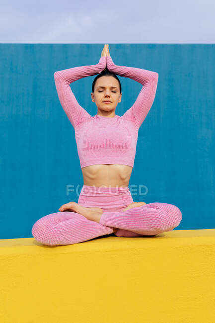 Cuerpo completo de hembra descalza en ropa deportiva sentada en pose de loto con los brazos elevados y los ojos cerrados - foto de stock