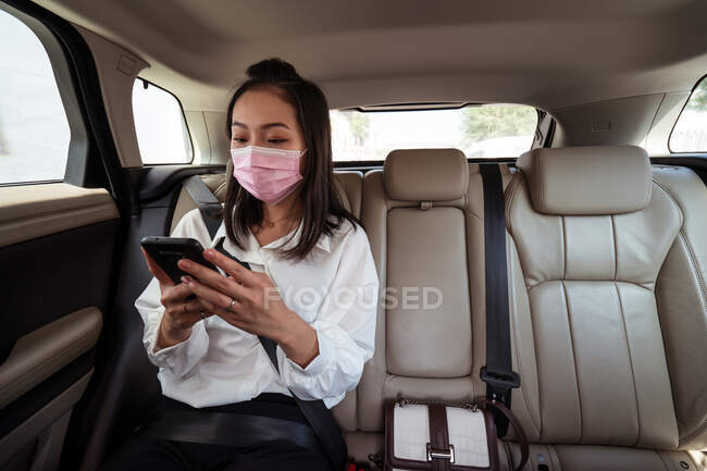 Ethnic female passenger with fastened seat belt using the cell phone while riding in protective mask on backseat in taxi — Stock Photo