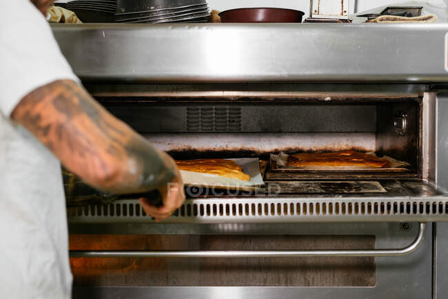 Crop faceless male baker with tattoos on arm baking cake in large metal oven during work in bakery — Stock Photo