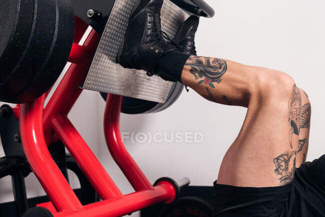 Vue latérale du sportif musclé avec des tatouages faisant des exercices sur la machine de presse de jambe pendant l'entraînement dans la salle de gym — Photo de stock