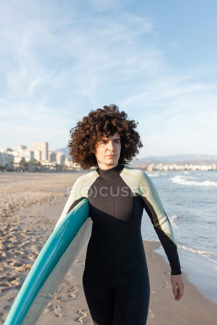 Junge, nachdenkliche Surferin im Neoprenanzug mit Surfbrett geht weg und blickt auf die Küste, die vom winkenden Meer angespült wird — Stockfoto