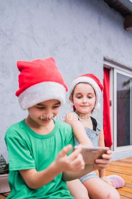 Діти в червоних капелюхах Санта переглядають мобільний телефон, сидячи в світлій кімнаті під час святкування свята — стокове фото