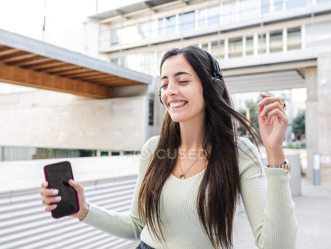 Fröhliche junge Frau mit langen braunen Haaren in lässiger Kleidung geht bei Tageslicht mit geschlossenen Augen durch die Stadt und hört Musik über Kopfhörer. — Stockfoto