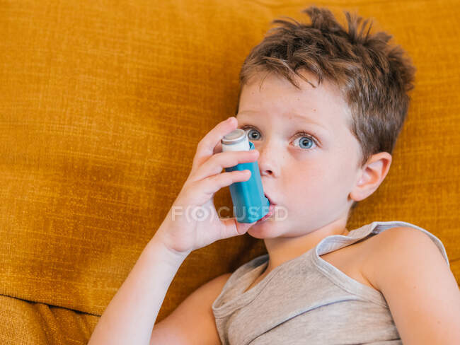 Vista lateral do menino com asma usando inalador enquanto sentado no sofá em casa — Fotografia de Stock