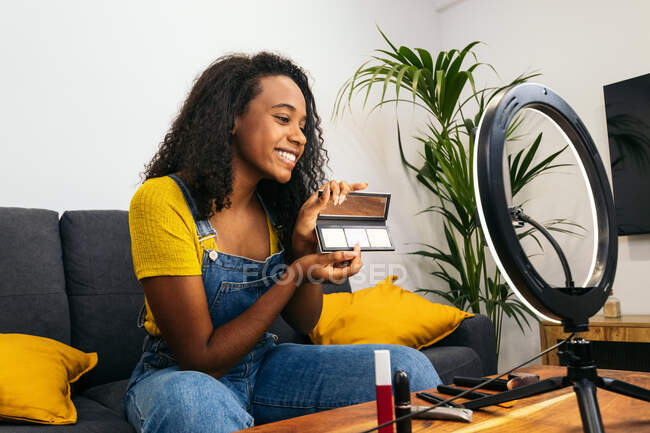 African American lady demonstrating powder palette during vlog recording via cellphone on LED ring lamp — Stock Photo