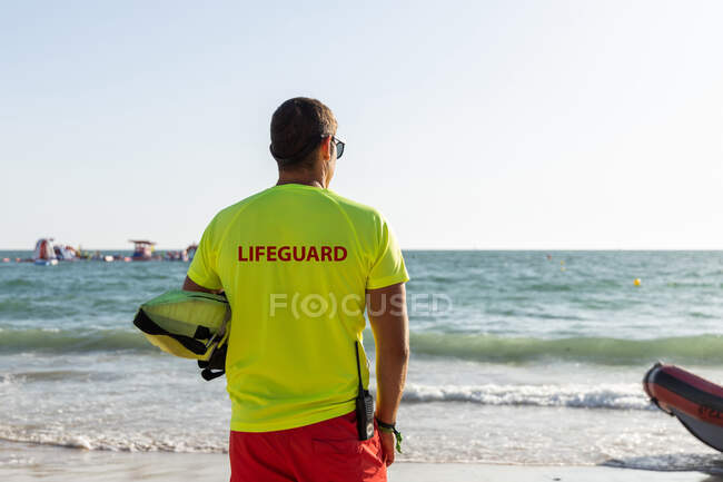 Back view of anonymous male lifeguard in shorts and t shirt and maintaining safety on sandy seashore — Stock Photo