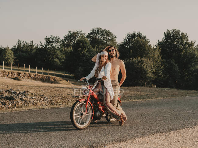 Corpo inteiro de casal hippie montando em vermelho ciclomotor na estrada de asfalto durante a viagem na natureza com árvores no dia de verão — Fotografia de Stock