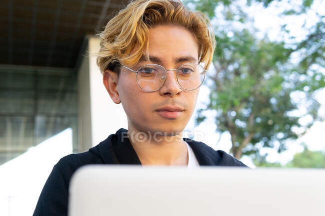 Concentrated young male freelancer typing on modern netbook while sitting on street with green trees in city during online work — Stock Photo