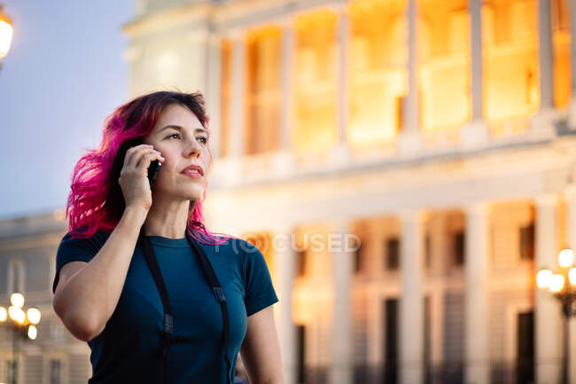 Cheerful female with pink hair phone calling while standing on street with streetlight near classic glowing building in city — Stock Photo