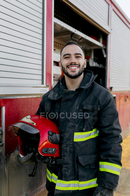 Cheerful bearded young firefighter standing near fire truck and holding red hardhat in hand while smiling and looking at camera — Stock Photo