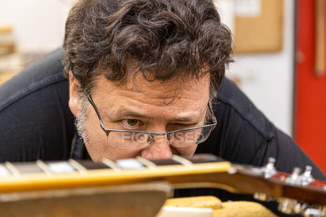 Cortar luthier masculino em roupas de trabalho e óculos ajustando porca branca no pescoço da guitarra enquanto trabalhava em oficina profissional com equipamentos — Fotografia de Stock
