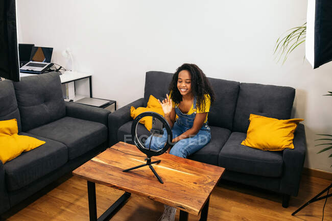 From above of ethnic woman sitting on couch and greeting audience while recording vlog for smartphone placed on ring lamp — Stock Photo