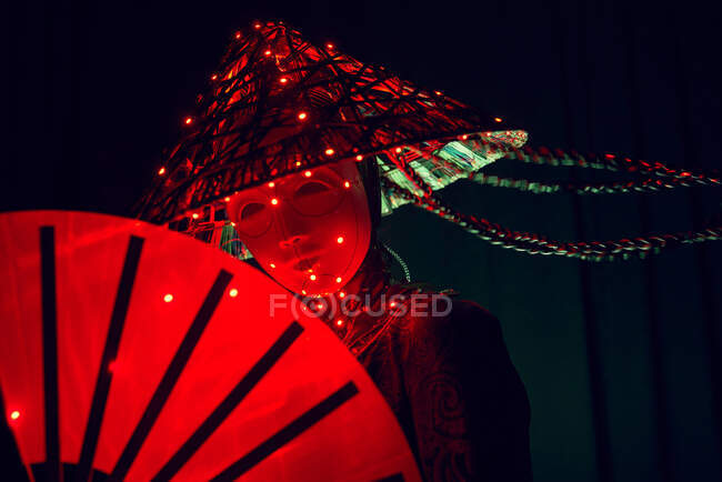 Unerkennbare Frau mit Maske in kreativem traditionellem Outfit und vietnamesischer Kopfbedeckung mit roter Beleuchtung im dunklen Studio auf schwarzem Hintergrund während des Auftritts — Stockfoto