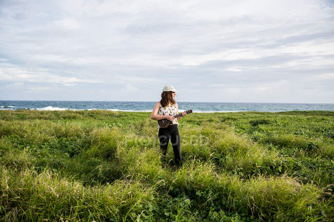 Calma músico femenino con cabello castaño en ropa casual de pie jugando en ukelele mientras mira hacia otro lado en el campo con hierba verde contra colinas a la luz del sol - foto de stock