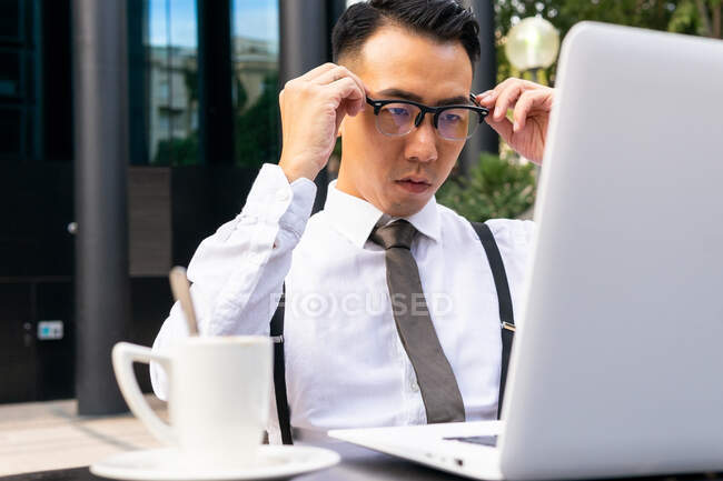 Gut gekleideter männlicher Jungunternehmer setzt Brille gegen Tisch mit Netbook und Heißgetränk in Straßencafeteria auf — Stockfoto