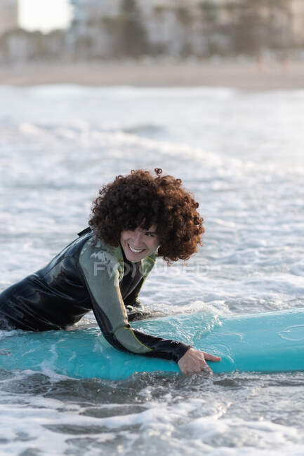 Вид сбоку на молодую счастливую женщину-серфингистку в гидрокостюме, лежащую на волнах моря и наслаждающуюся летним днем — стоковое фото