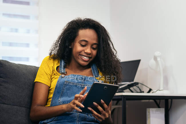 Mujer afroamericana positiva en traje de mezclilla sentada en el sofá y sonriendo mientras navega por la tableta en casa - foto de stock
