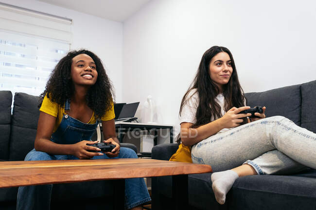 Amigos femininos multirraciais positivos sentados no sofá e jogando videogame enquanto passam tempo juntos em casa — Fotografia de Stock