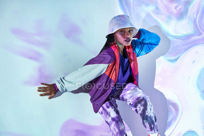 From above young Dominican female teenager in trendy outfit and hat sitting near white wall with creative abstract projections and looking at camera — Stock Photo