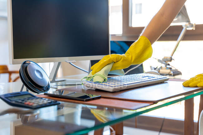 Crop governante femminile anonimo in guanti di gomma gialla pulire polvere dalla tastiera del computer moderno posto su un tavolo di vetro in una spaziosa sala da lavoro — Foto stock