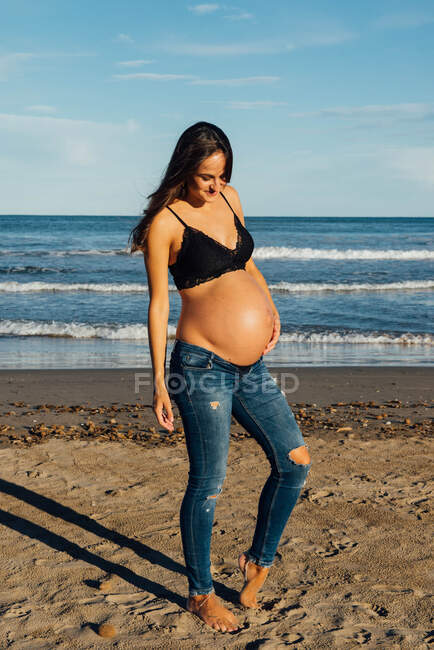 Donna incinta in reggiseno abbracciando delicatamente la pancia nuda mentre in piedi contro il mare ondulato e guardando giù con sorriso nella giornata di sole ventoso — Foto stock