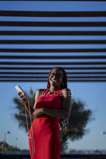 Von unten positiver Afroamerikanerin mit Zöpfen im trendigen Outfit, die mit Smartphone in der Hand auf der Straße steht und bei sonnigem Wetter lacht — Stockfoto