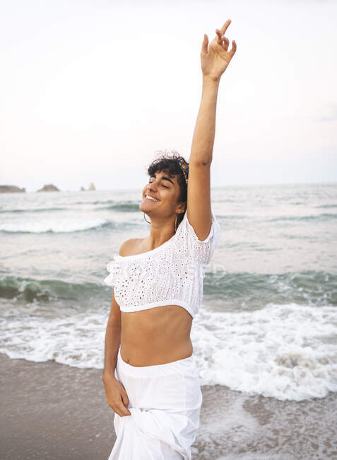 Positive ethnische Frau in weißem, stylischem Outfit und mit lockigem Haar am Strand stehend und mit geschlossenen Augen lachend, während er den Arm hebt — Stockfoto