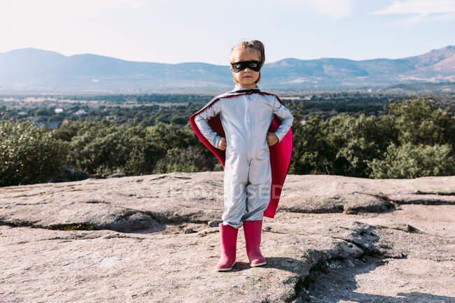 Corpo inteiro de menina pequena em traje de super-herói com as mãos na cintura em pé na colina rochosa — Fotografia de Stock