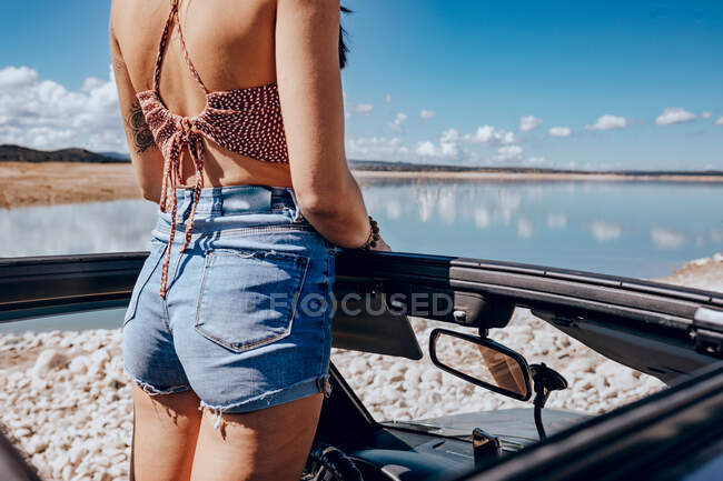 Back view of unrecognizable young tattooed female traveler in top and denim shorts standing in convertible SUV car parked on sandy beach and admiring picturesque seascape under cloudless blue sky — Stock Photo