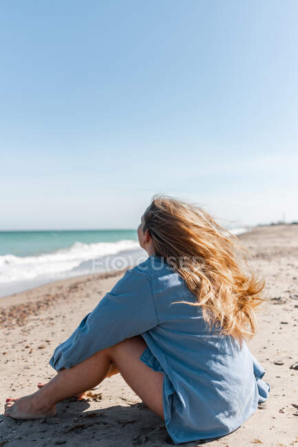 Donna irriconoscibile in camicia seduta sulla spiaggia sabbiosa vicino al mare mentre si gode la giornata estiva — Foto stock
