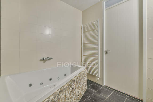 Diseño interior de baño moderno con pared de baldosas beige y suelo gris y toalla blanca calentada riel y bañera con efecto masaje y puerta blanca - foto de stock