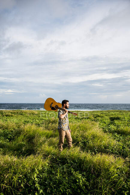 Calm man musician in casual clothes standing with acoustic guitar on shoulder among green grass on coast of ocean in summertime in daylight — Stock Photo