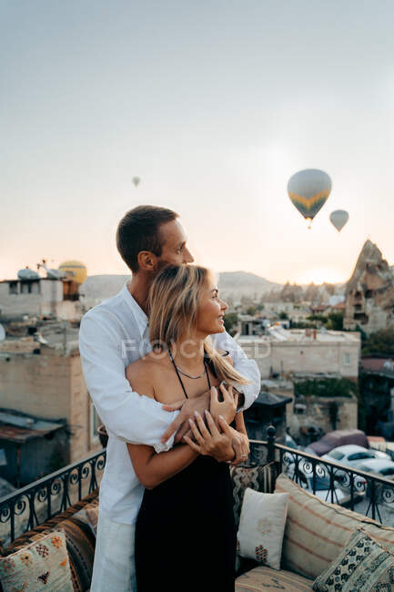 Vue latérale de l'homme aimant embrassant la femme par derrière regardant loin sur la terrasse du toit avec des montgolfières dans le ciel du soir en Cappadoce Turquie — Photo de stock