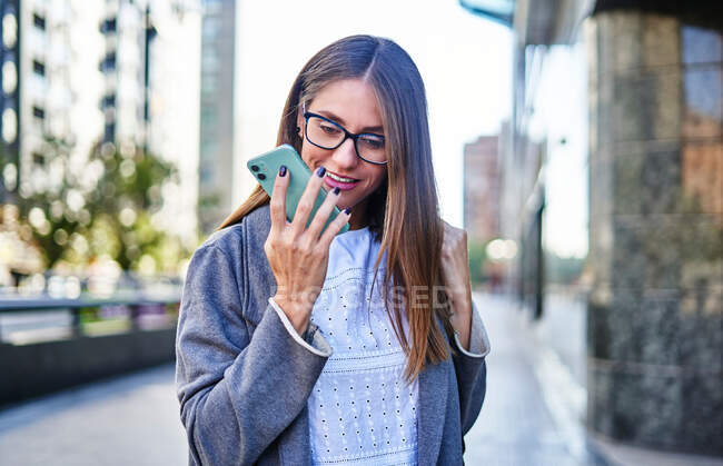 Positive female with long straight fair hair in eyeglasses and formal wear recording voice message while standing on street — Stock Photo