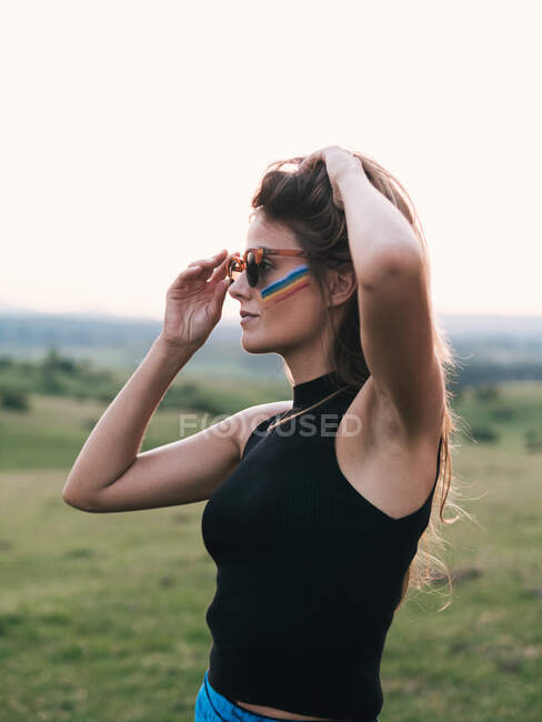 Gros plan d'une femme avec un arc-en-ciel sur la joue et les mains tenant lunettes et cheveux — Photo de stock