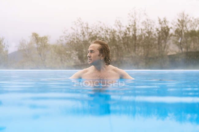 Homem calmo sem camisa com cabelos encaracolados nadando na piscina com água quente e olhando para longe contra árvores que crescem na natureza na Islândia durante o dia — Fotografia de Stock