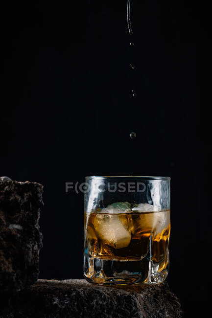 Gocce di whisky che cadono su cubetti di ghiaccio servite in vetro cristallo posto su una superficie ruvida su sfondo nero — Foto stock
