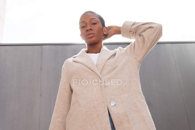 Sério backlit afro-americano fêmea com cabelo curto olhando para a câmera enquanto estava perto da parede na rua com luz solar brilhante, enquanto ele toca sua cabeça com a mão — Fotografia de Stock