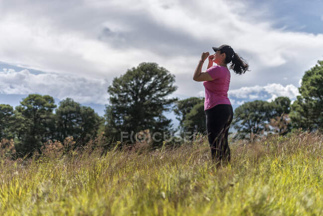 Вид збоку самка з піднятими руками, що стоять на трав'янистому полі з зеленими деревами, насолоджуючись природою під час піших прогулянок — стокове фото