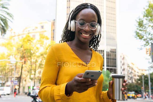 Encantada mujer afroamericana con taza termo escuchando música en auriculares mientras mensajería de texto en el teléfono celular en la calle en la ciudad - foto de stock