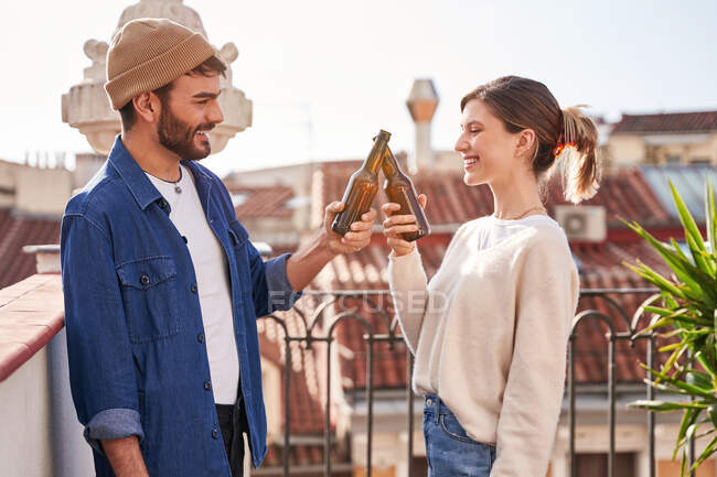 Очарованные юные друзья в повседневной одежде, звенящие бутылки пива, расслабляясь на террасе вместе — стоковое фото