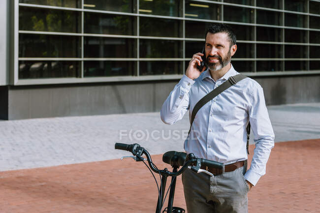 Male worker in formal wear standing with bicycle and having conversation on mobile phone in downtown — Stock Photo