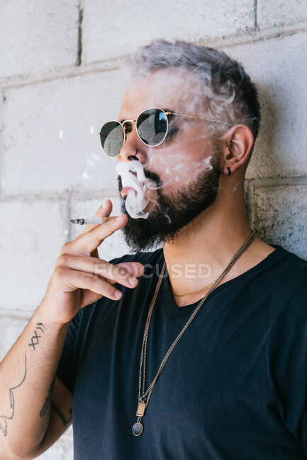 Thoughtful male with beard and mustache in casual clothes and sunglasses standing near grey wall of building and smoking cigarette in daylight — Stock Photo