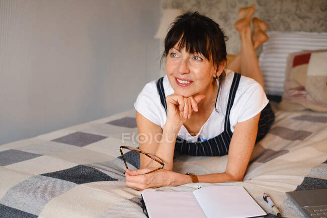 Positive middle age female leaning on hand while lying on bed with opened notepad and eyeglasses and looking away — Stock Photo