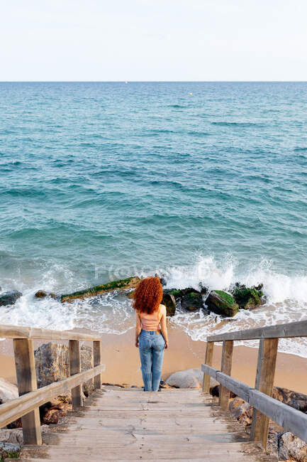 High angle back view of unrecognizable female traveler with long curly ginger hair standing on wet sandy beach washed by foamy splashing waves — Stock Photo