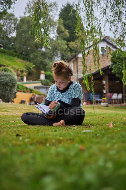 Cuerpo completo de chica descalza concentrada leyendo interesante libro mientras está sentado en el césped cubierto de hierba en el patio trasero contra el edificio residencial en el campo - foto de stock