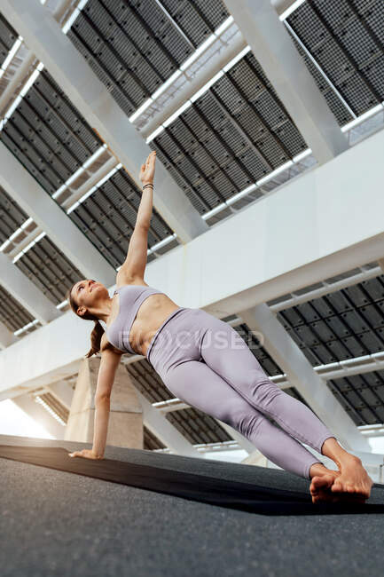 Donna scalza in abbigliamento sportivo pratica postura Utthita Vasishthasana sul tappeto vicino al pannello fotovoltaico sulla strada durante un intenso allenamento yoga — Foto stock