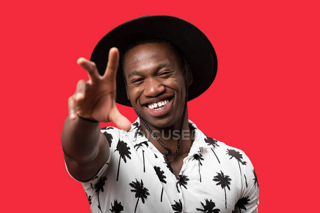 Allegro maschio afroamericano in elegante cappello e camicia alla moda ridendo felicemente e raggiungendo mano alla macchina fotografica contro lo sfondo rosso — Foto stock